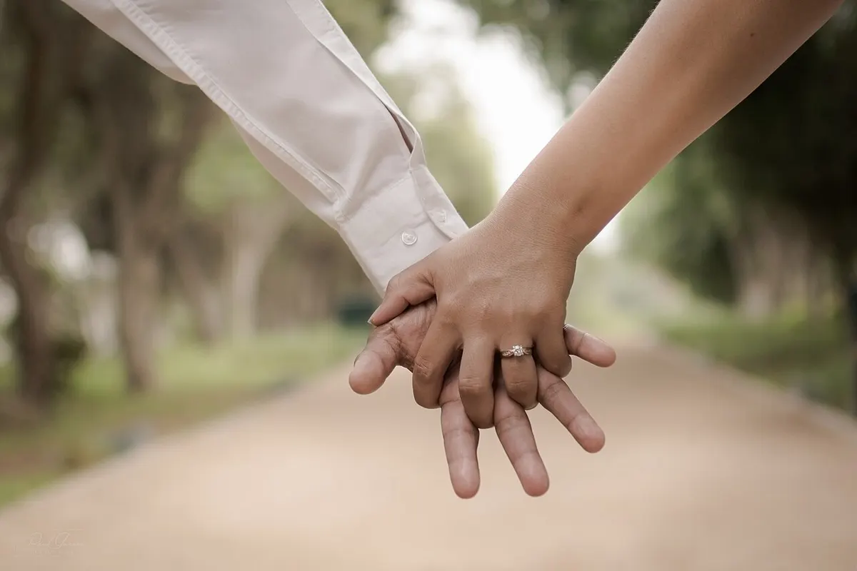 Dos manos entrelazadas con un anillo de compromiso en un camino entre árboles